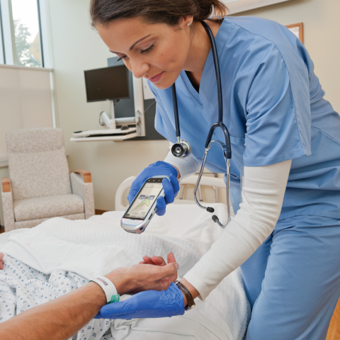 Pflegefachperson mit einem mobilen Terminal scannt den Barcode auf dem Patientenarmband am Bett eines Patienten