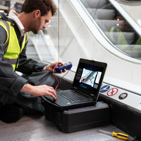 Service-Techniker im Ausseneinsatz verwendet ein robustes Notebook und verbundene Messgeräte bei der Wartung einer Rolltreppe