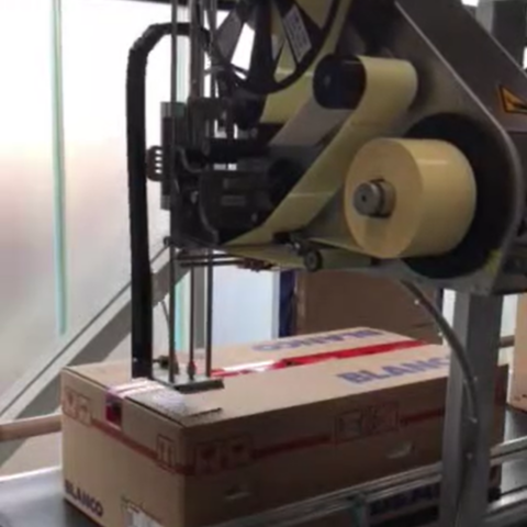 Automatischer Etikettenapplikator klebt ein Versandetikett auf eine Kartonschachtel an einer Verpackungslinie